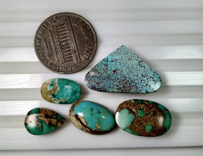 Puede incluir: Una colecci&oacute;n de piedras preciosas de turquesa en varias formas, incluyendo ovaladas y en forma de l&aacute;grima, con patrones de matriz marr&oacute;n. Se incluye una moneda de un centavo estadounidense para referencia de tama&ntilde;o. Las piedras est&aacute;n sobre una superficie blanca.