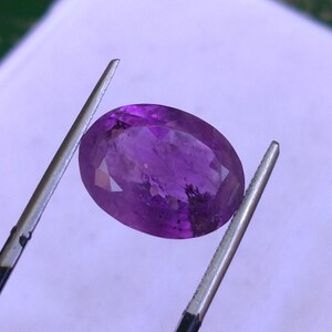 May include: A faceted, oval-shaped amethyst gemstone, held by tweezers. The gem displays a deep purple hue with internal inclusions. The tweezers are silver-colored, and the background is a soft, blurred white.