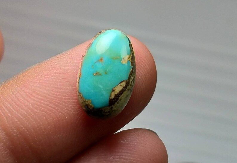 Puede incluir: Una piedra preciosa ovalada de turquesa con una superficie pulida. La piedra presenta un vibrante tono azul-verdoso con patrones de matriz marr&oacute;n y beige en los bordes. La piedra preciosa se sostiene entre los dedos.