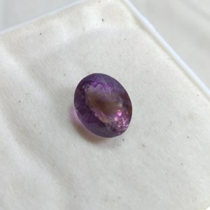 May include: An oval-shaped gemstone with a gradient of colors, primarily purple and a hint of brown. The polished surface reflects light, showcasing the stone's internal structure. The gemstone is set against a white background.