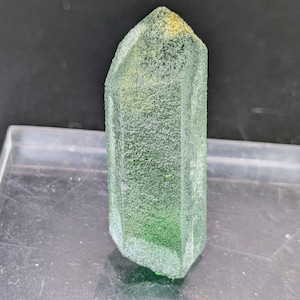 Puede incluir: Un cristal verde facetado con una superficie texturizada. El cristal es de color verde claro con una punta ligeramente amarilla. El cristal está de pie y tiene una forma geométrica.