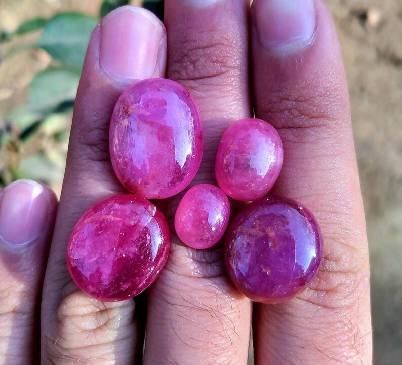Peut inclure: Six pierres pr&eacute;cieuses rubis polies, de forme ovale et de tailles vari&eacute;es. Les rubis pr&eacute;sentent une teinte rouge-rose profonde et vibrante, avec une surface brillante et r&eacute;fl&eacute;chissante. Les pierres sont tenues dans une main.
