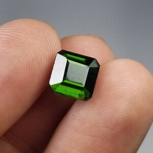 Pode incluir: Uma vibrante gema verde esmeralda com um corte retangular. A gema &eacute; segurada entre os dedos, mostrando suas facetas polidas e sua cor profunda. A clareza e o brilho da gema s&atilde;o destacados em um fundo neutro.