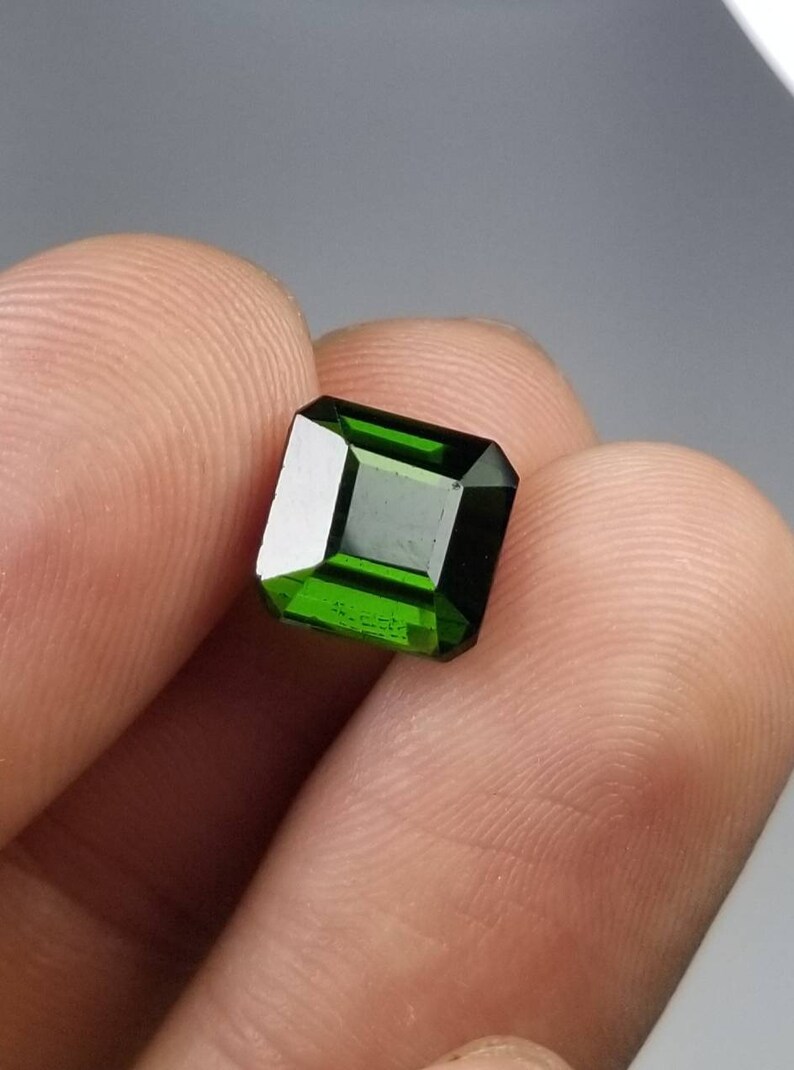 Pode incluir: Uma vibrante gema verde esmeralda com um corte retangular. A gema &eacute; segurada entre os dedos, mostrando suas facetas polidas e sua cor profunda. A clareza e o brilho da gema s&atilde;o destacados em um fundo neutro.