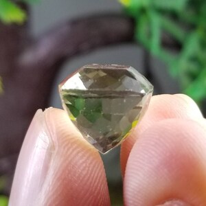 May include: A faceted, olive-green gemstone with a diamond-like cut. The gem is held between two fingers, showcasing its clarity and multiple reflective surfaces. The background is blurred, with hints of green foliage.