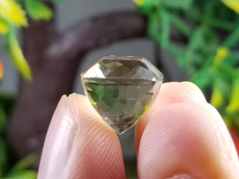 May include: A faceted, olive-green gemstone with a diamond-like cut. The gem is held between two fingers, showcasing its clarity and multiple reflective surfaces. The background is blurred, with hints of green foliage.