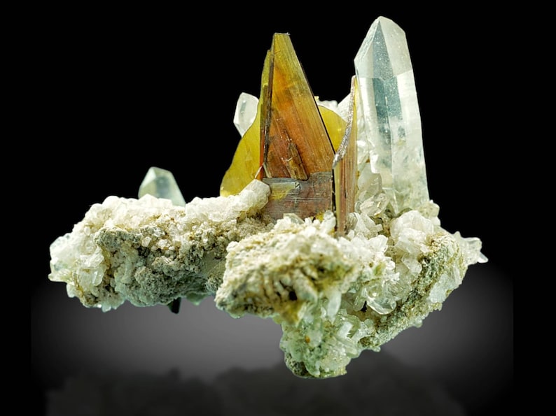 May include: A cluster of crystals featuring a central, multi-faceted, brown and yellow crystal formation. Clear, pointed quartz crystals and smaller, white crystals are also present. The crystals rest on a base of light green and white mineral deposits, against a black background.