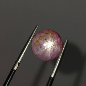 May include: A spherical, polished gemstone, held by tweezers. The gem is a pale pink colour with golden inclusions and a star-like effect. The surface has fine lines. The background is a solid grey.