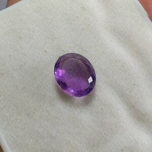May include: A faceted, oval-shaped amethyst gemstone. The gem is a deep purple color with a polished surface, reflecting light. The gemstone is set against a white background.