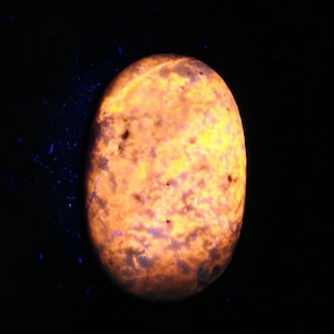 Könnte beinhalten: Ein ovaler, leuchtender Edelstein mit einer feurigen Farbpalette aus Orange und Gelb. Der Stein hat dunkelbraune und schwarze Einschlüsse und ist vor einem schwarzen Hintergrund platziert. Der Edelstein scheint von innen beleuchtet zu sein.