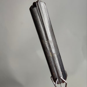 May include: A dark, elongated black tourmaline crystal with vertical striations. The crystal is held by a silver metal stand, and the background is a soft grey. The top of the crystal has a rough, uneven texture.