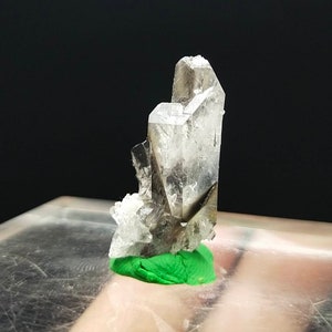 May include: A clear quartz crystal cluster with smoky inclusions. The crystal is mounted on a green base, showcasing its natural form and clarity. The quartz has a multifaceted structure, reflecting light and displaying a range of tones.
