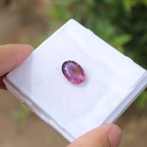May include: An oval-shaped gemstone with a gradient of pink and purple hues. The gem is set on a white surface, showcasing its polished facets. The background is blurred, suggesting an outdoor setting, highlighting the gem's vibrant colors.