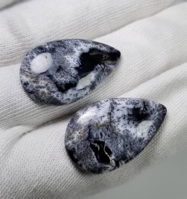 Pode incluir: Duas pedras preciosas em forma de gota com um padr&atilde;o marcante em preto e branco. As pedras polidas exibem um efeito marmorizado, com &aacute;reas de preto e branco s&oacute;lido intercaladas com detalhes cinza e salpicados. As pedras est&atilde;o sobre um fundo de tecido branco.