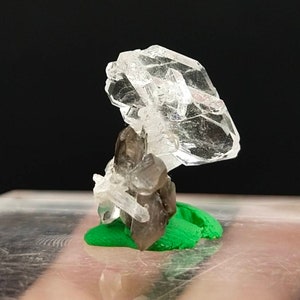 Puede incluir: Un grupo de cristales de cuarzo transparente con un cristal grande y plano en la parte superior y cristales de cuarzo ahumado más pequeños debajo. Los cristales están colocados sobre una pequeña base verde brillante. El fondo es negro.