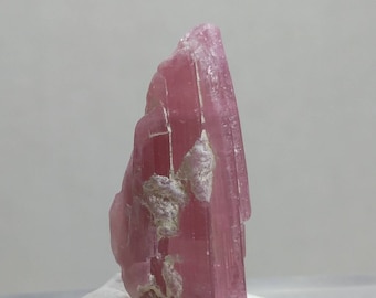 May include: A raw, pink tourmaline crystal with a rough, textured surface. The crystal has a vertical, elongated shape with a few white patches. The color is a vibrant, saturated pink, and the crystal is displayed against a neutral background.