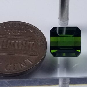 Pode incluir: Uma pedra preciosa verde escura, lapidada em esmeralda, &eacute; exibida ao lado de uma moeda de um centavo americana. A pedra preciosa tem formato retangular com cantos cortados. A moeda de um centavo &eacute; uma moeda de cor cobre.