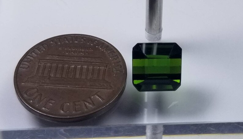 Pode incluir: Uma pedra preciosa verde escura, lapidada em esmeralda, &eacute; exibida ao lado de uma moeda de um centavo americana. A pedra preciosa tem formato retangular com cantos cortados. A moeda de um centavo &eacute; uma moeda de cor cobre.