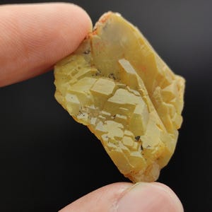 Op de afbeelding: Een close-up van een geelgroen mineraalmonster met een kristallijne structuur. Het mineraal wordt tussen twee vingers gehouden tegen een zwarte achtergrond. Het monster heeft een ruw, getextureerd oppervlak met verschillende geometrische vormen.
