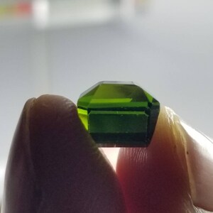Pode incluir: Uma gema verde esmeralda facetada com formato octogonal. A gema &eacute; segurada entre dois dedos, mostrando sua clareza e cor vibrante. As facetas da gema refletem a luz, destacando seu corte e brilho.