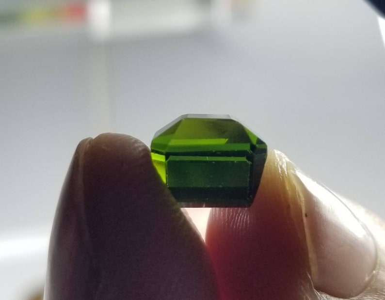 Pode incluir: Uma gema verde esmeralda facetada com formato octogonal. A gema &eacute; segurada entre dois dedos, mostrando sua clareza e cor vibrante. As facetas da gema refletem a luz, destacando seu corte e brilho.
