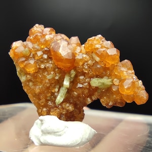 Pode incluir: Um aglomerado de cristais castanho-alaranjados com uma textura granular brilhante. Vários cristais alongados verde-claro estão espalhados por todo o aglomerado. O espécime mineral é montado numa pequena base branca, mostrando a sua beleza natural.