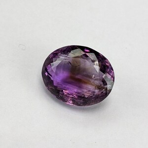 May include: An oval-shaped amethyst gemstone with a faceted cut. The gem displays a gradient of colors, from deep purple to lighter shades, with hints of brown. The stone is set against a white background.