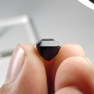 Pode incluir: Uma pedra preciosa preta facetada, segurada entre os dedos. A gema tem uma forma retangular. A pedra &eacute; de uma cor preta profunda e opaca, e as bordas s&atilde;o n&iacute;tidas e bem definidas. A gema est&aacute; sendo segurada contra a luz.