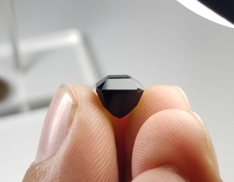 Pode incluir: Uma pedra preciosa preta facetada, segurada entre os dedos. A gema tem uma forma retangular. A pedra &eacute; de uma cor preta profunda e opaca, e as bordas s&atilde;o n&iacute;tidas e bem definidas. A gema est&aacute; sendo segurada contra a luz.