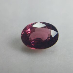 May include: A faceted, oval-cut gemstone in a deep magenta colour. The gem reflects light, showing its cut and clarity. The stone is set against a plain white background, highlighting its colour and form.