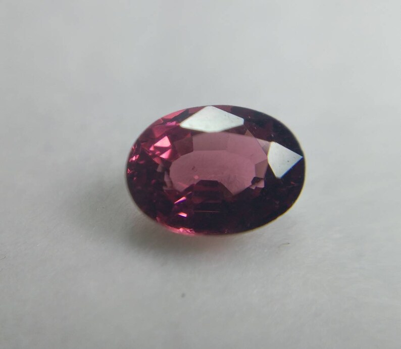 May include: A faceted, oval-cut gemstone in a deep magenta colour. The gem reflects light, showing its cut and clarity. The stone is set against a plain white background, highlighting its colour and form.