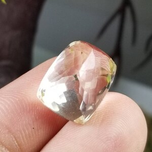 May include: A faceted, rectangular gemstone with a pale yellow hue. The gem is held between fingers, showcasing its polished surfaces and internal clarity. The background features blurred greenery.
