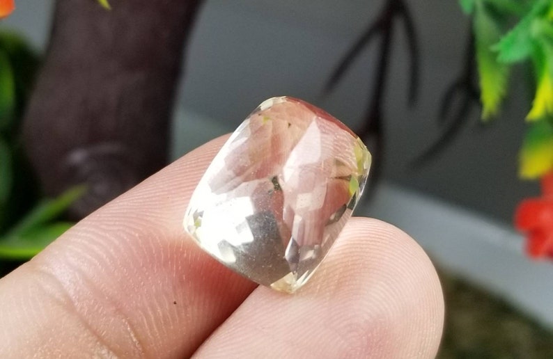 May include: A faceted, rectangular gemstone with a pale yellow hue. The gem is held between fingers, showcasing its polished surfaces and internal clarity. The background features blurred greenery.