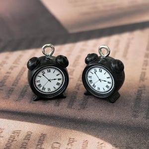 May include: Two small, black alarm clock charms with white clock faces and Roman numerals. The clocks have a silver loop at the top for attaching to jewelry or other crafts. The hands of the clocks are pointing to different times.