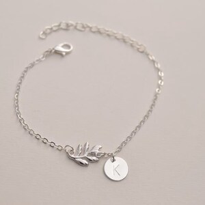 May include: A silver chain bracelet featuring a delicate leaf charm and a small round charm engraved with the letter 'K'.