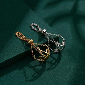 May include: Two pendant necklaces, one gold-toned and one silver-toned, rest on a dark green surface. Each necklace features a chain that leads to a decorative openwork pendant with a geometric design. The pendants are suspended from a loop.