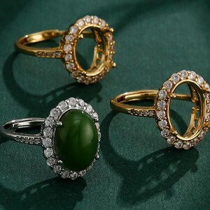 May include: Three rings on a dark green surface. Two are gold-toned, with a halo of small, clear stones around the oval setting. One ring has a silver-toned band and a dark green oval stone surrounded by a halo of clear stones.