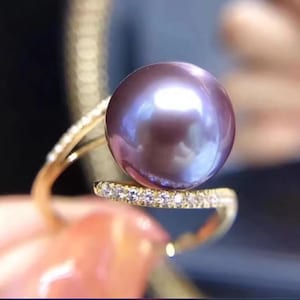 May include: A close-up of a gold ring featuring a large, iridescent purple pearl. The ring band is adorned with small, sparkling diamonds. The pearl has a smooth, reflective surface, and the ring is set against a blurred background.