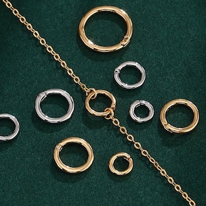 May include: An assortment of gold and silver-colored jewelry clasps and a gold chain are arranged on a dark green surface. The clasps are circular, with a hinged opening. The chain is delicate, with small links. The jewelry components are displayed in a top-down view.