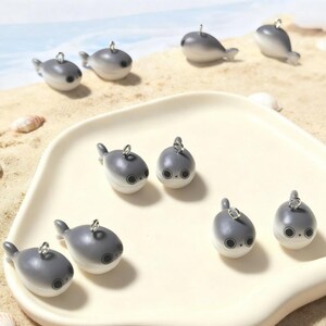 May include: A collection of gray and white whale-shaped charms with silver-colored loops for attaching to jewelry or other crafts. The charms are arranged on a white tray and a sandy surface, with a blurred beach scene in the background.