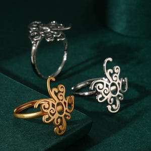 May include: Three rings with ornate, openwork designs are displayed against a dark green background. One ring is gold-toned, while the other two are silver-toned. The rings feature swirling, floral-like patterns.