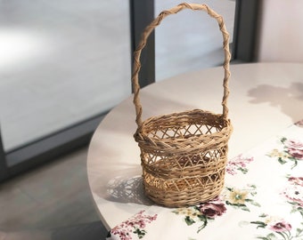 Könnte beinhalten: Ein geflochtener Korb aus Weide mit einem Griff. Der Korb ist hellbraun und hat ein geflochtenes Muster. Der Korb steht auf einem weißen Tisch mit einer floralen Tischdecke.