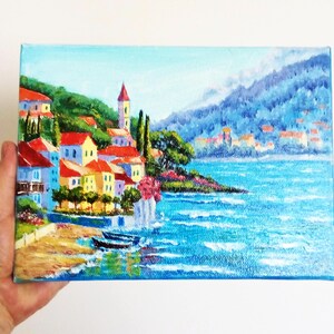 May include: A painting depicting a coastal town with colourful buildings, a church steeple, and a blue sea with whitecaps. The town is nestled on a hillside with green trees and a mountain in the background.