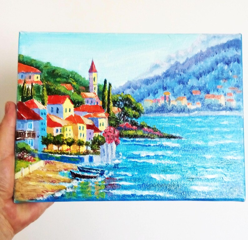 May include: A painting depicting a coastal town with colourful buildings, a church steeple, and a blue sea with whitecaps. The town is nestled on a hillside with green trees and a mountain in the background.