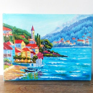 May include: A painting depicting a colourful seaside town with a blue sky and ocean. The town is built on a hillside with a church steeple in the centre. The ocean is a bright blue with whitecaps.