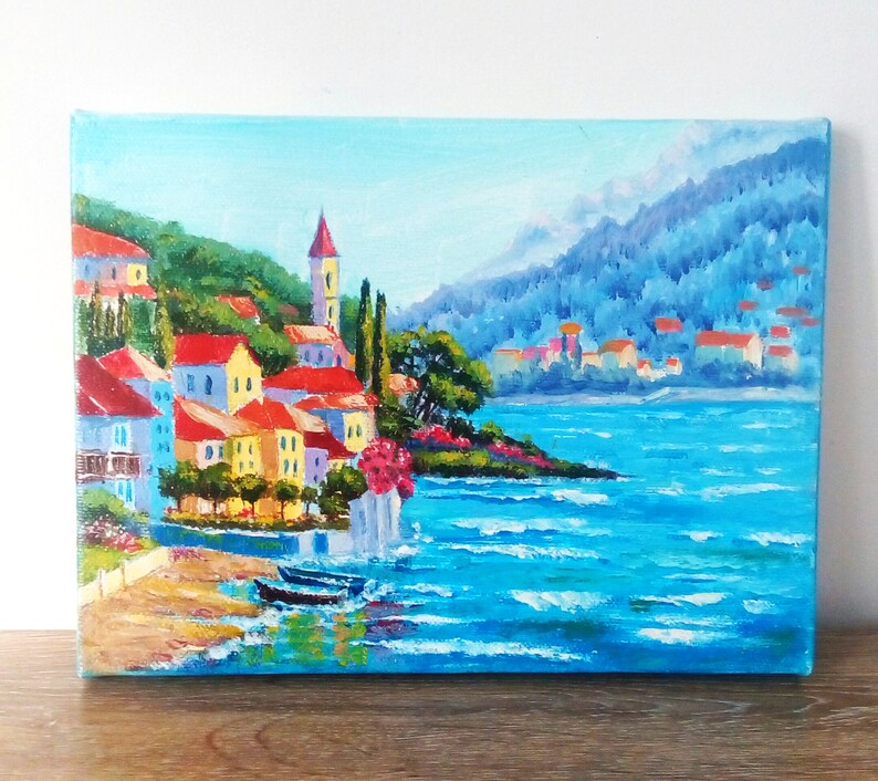 May include: A painting depicting a colourful seaside town with a blue sky and ocean. The town is built on a hillside with a church steeple in the centre. The ocean is a bright blue with whitecaps.