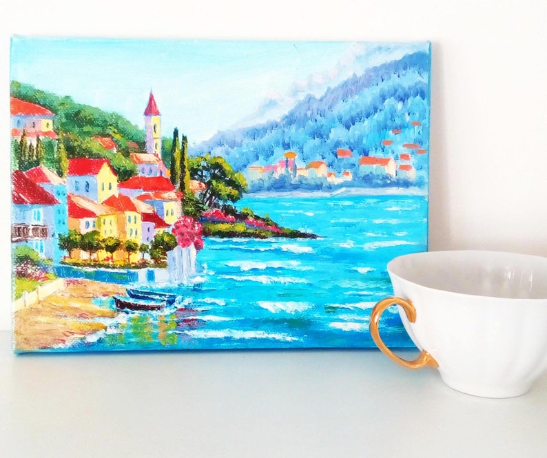 May include: A painting depicting a coastal town with colourful buildings, a church spire, and a blue sea with whitecaps. The town is nestled on a hillside with green trees and a mountain in the background.