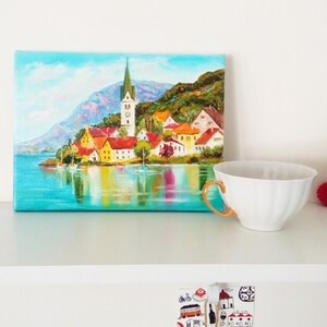 May include: A painting depicting a small town with a church spire, surrounded by water and trees. The painting is executed in a bright, colourful style.
