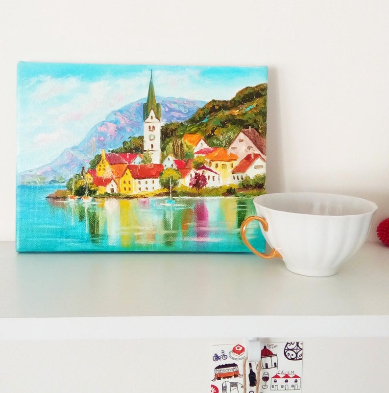 May include: A painting depicting a small town with a church spire, surrounded by water and trees. The painting is executed in a bright, colourful style.