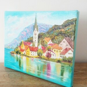 May include: A painting depicting a quaint village with a church spire, nestled in a valley with mountains in the background. The village is located on a lake with colourful reflections of the buildings in the water.